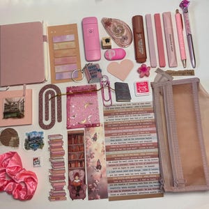 Könnte beinhalten: Eine Sammlung rosa Schreibwaren und Accessoires, darunter ein Notizbuch, Stifte, Aufkleber, ein Scrunchie und ein Federmäppchen. Die Artikel sind auf einer weißen Oberfläche angeordnet und bilden eine ansprechende Auswahl für das Journaling und kreative Projekte.