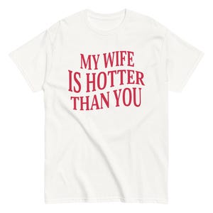 Może przedstawiać: Biała koszulka z czerwonym napisem "MY WIFE IS HOTTER THAN YOU". Koszulka ma okrągły dekolt i krótkie rękawy. Tekst jest pogrubiony, lekko zniekształcony.