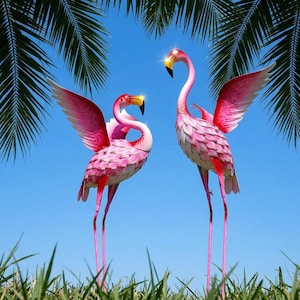 May include: Two pink metal flamingo sculptures with layered feather details and yellow beaks. Both flamingos are depicted with wings spread wide against a clear blue sky, framed by green palm fronds at the top and green grass at the bottom.