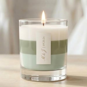 May include: A lit, layered candle in a clear glass. The candle has three horizontal sections: white, green, and light blue. A label with "M & J | 09.20.26" is affixed to the front. The candle sits on a light-coloured wooden surface.