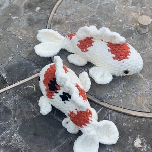 Puede incluir: Dos peces koi de ganchillo hechos a mano con hilo blanco suave. Un pez presenta manchas naranjas, mientras que el otro muestra una combinación de manchas naranjas y negras. Ambos peluches tienen ojos pequeños y oscuros y aletas detalladas.
