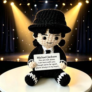 Könnte beinhalten: Gehäkelte Puppe von Michael Jackson in einem schwarzen Anzug, Hut und weißem Hemd. Die Puppe hält ein Schild mit dem Text "Michael Jackson, You are not alone. I am here with you. Though we're far apart, you're always in my heart."
