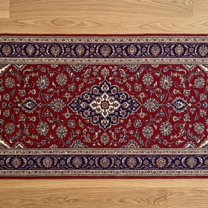 May include: A rectangular red rug with an intricate floral pattern and a navy blue border. The rug has white fringe on both ends and is placed on a wooden floor. The rug's design features a central medallion and repeating floral motifs.
