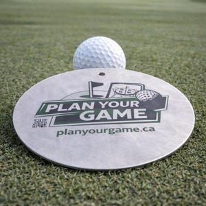 Peut inclure: Une balle de golf repose sur un marqueur circulaire argenté avec le texte "PLAN YOUR GAME" en vert et noir. Le marqueur présente également un graphique de terrain de golf et l'adresse du site Web, planurgame.ca. La scène se déroule sur un parcours de golf vert.