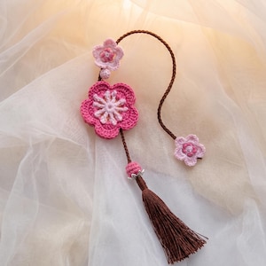 May include: A handmade crochet floral ornament featuring three pink flowers of varying sizes, connected by a brown cord. A brown tassel hangs from the largest flower. The ornament is set against a soft, off-white background.