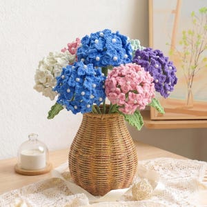May include: A bouquet of handmade crochet hydrangeas in a woven wicker vase. The flowers are in shades of blue, pink, purple, and white, with pearl accents. The vase is brown and sits on a wooden surface.