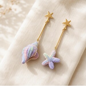 May include: A pair of drop earrings featuring gold-tone star studs attached to thin gold-tone bars. One earring displays a crocheted seashell in pastel variegated colors, while the other features a crocheted starfish in matching pastel hues.