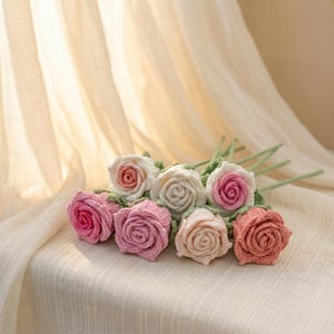 Può includere: Un gruppo di rose all'uncinetto fatte a mano in varie tonalità di rosa e bianco, con steli e foglie verdi. I fiori sono disposti su una superficie color crema, con uno sfondo morbido e neutro.