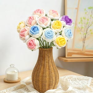May include: A bouquet of colorful, crocheted roses in a woven wicker vase. The flowers are in shades of pink, yellow, blue, and purple, with green stems. The vase is brown and sits on a wooden table with a lace runner.