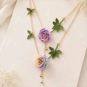 May include: Two gold chain necklaces with crocheted flower pendants. One pendant is a gradient of pastel colors, the other is purple. Both necklaces have gold bead accents and green leaf details.