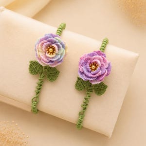 May include: Two handmade crochet flower brooches. Each brooch features a multi-colored rose with green leaves and stem. The roses have shades of purple, pink, and yellow. The stems are a twisted green yarn.