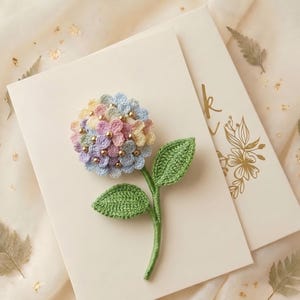 May include: A handmade crochet hydrangea flower in pastel colors of blue, pink, yellow, and purple, with gold beads. The flower sits on a white card with a green stem and leaves. A second card with gold floral design is behind it.