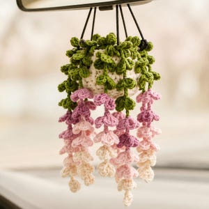 May include: A crocheted hanging plant ornament featuring a small white pot with green vines and cascading flowers in shades of purple, pink, and cream. The plant is suspended by black strings, designed to hang from a vehicle rearview mirror.