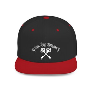 Op de afbeelding: Zwarte en rode baseballcap met de tekst "Prison Dog Locksmith" in witte letters. De cap heeft een platte klep en een rode knop. Het ontwerp bevat gekruiste sleutels.
