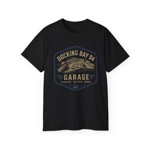 May include: A black short-sleeved t-shirt featuring a graphic of a spaceship inside a blue and beige hexagonal emblem. The text reads DOCKING BAY 94 GARAGE SERVICE REPAIR MODS with a small Rebel Alliance symbol at the bottom of the design.