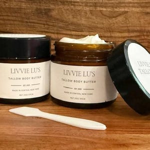 May include: Two glass jars of Livvie Lu's Tallow Body Butter with black lids and white labels. One jar is open, revealing the product. A white spatula lies in front. The text on the label reads "Tallow Body Butter" and "Made in Central New York".