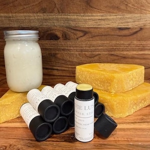 Könnte beinhalten: Ein klares Glasgefäß mit weißer Creme, mehrere schwarze zylindrische Butterstifte mit der Aufschrift Vie Lu's Butter Stick und zwei große Blöcke aus gelbem Bienenwachs. Ein Butterstift steht aufrecht ohne seine schwarze Kappe.
