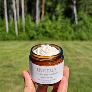 Puede incluir: Un pequeño frasco de vidrio ámbar lleno de manteca corporal de sebo batida blanca. La etiqueta blanca presenta el texto LIVVIE LU'S TALLOW BODY BUTTER, EST. 2025, MADE IN CENTRAL NEW YORK, y NET 2 OZ/ 57 G. El frasco tiene una tapa negra.