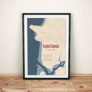 Peut inclure: Impression d'art encadrée représentant une carte de Croyde & Saunton, Devon, Angleterre. L'œuvre utilise une palette de couleurs beige, bleu et marron. Le texte "Croyde & Saunton" est en rouge. L'impression est dans un cadre noir, sur un sol en bois.