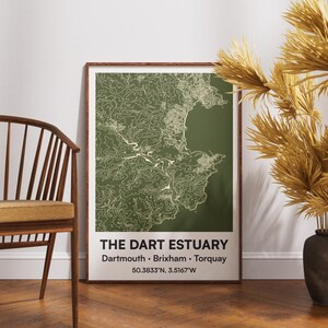 Puede incluir: Un póster enmarcado del estuario de Dart, con un mapa topográfico detallado en verde y blanco. El póster incluye el texto "The Dart Estuary" y coordenadas. Una silla de madera y un jarrón decorativo con plantas secas están en primer plano.