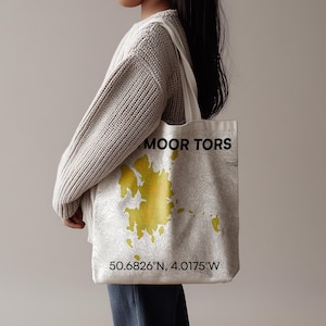 Könnte beinhalten: Cremefarbene Tragetasche mit gelb-weißem topografischem Kartendesign. Auf der Tasche steht "MOOR TORS" und die Koordinaten 50.6826°N, 4.0175°W. Eine Person hält die Tasche und trägt einen cremefarbenen Pullover.