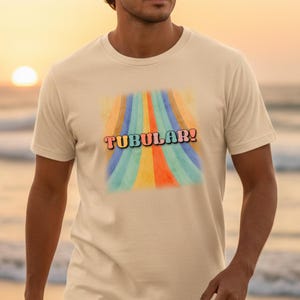 Op de afbeelding: Een crèmekleurig T-shirt met het woord "TUBULAR!" in een retro lettertype. De afbeelding heeft een kleurrijk, regenboogachtig ontwerp. Het shirt heeft korte mouwen en is gemaakt van een zacht materiaal.