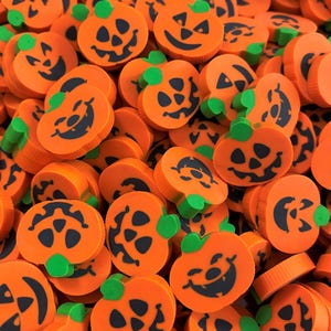 May include: A collection of small, round, orange pumpkin-shaped erasers. Each eraser features a black printed jack-o'-lantern face with triangular eyes and a jagged mouth, topped with a small, bright green stem protruding from the top edge.