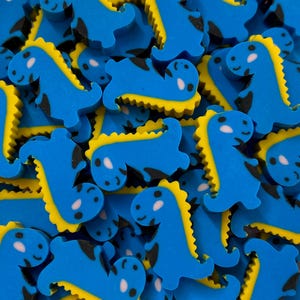 May include: A collection of small, blue dinosaur-shaped erasers featuring a bright yellow serrated spine, black accents on the body, and a white patch on the face. Each individual piece is molded into a cartoon-like standing or lying posture.
