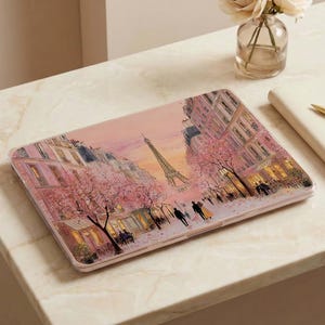 Peut inclure: Une coque rigide pour ordinateur portable présentant une illustration imprimée d'une rue parisienne avec la tour Eiffel en arrière-plan, des cerisiers en fleurs roses et des silhouettes marchant sur le trottoir aux tons doux et chauds.