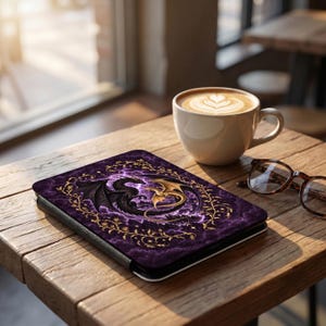 May include: A rectangular e-reader case featuring a purple background with a dark dragon and a gold dragon intertwined in a circular formation, surrounded by a gold vine border. The case is placed on a wooden surface next to a coffee cup and glasses.