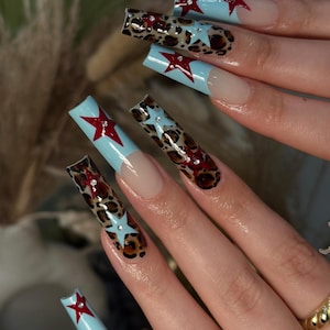 May include: Long, square artificial nails with a light blue base, red star accents, and leopard print designs. Some nails feature clear star-shaped rhinestones. The nails have a natural-looking, nude-coloured base near the cuticle.