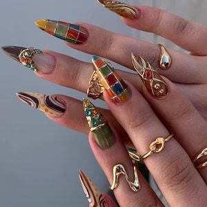 Puede incluir: Una mano que muestra un conjunto de uñas acrílicas largas y puntiagudas. Las uñas presentan varios diseños, incluyendo un patrón de tablero de ajedrez colorido, adornos dorados y remolinos abstractos en tonos marrones y blancos. Varios anillos dorados adornan los dedos.