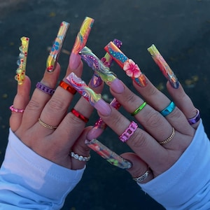 May include: A close-up of long, colorful acrylic nails with various designs, including floral patterns and abstract swirls. The nails are adorned with multiple rings in various colors, including pink, red, green, and blue. The nails are a vibrant display of nail art.