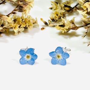 May include: Two rings with light blue flower designs. The flowers have a yellow center and are set on silver-colored bands. Dried yellow flowers are in the background, creating a natural and delicate aesthetic. The rings are displayed on a white surface.