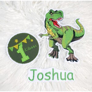 Könnte beinhalten: Gestickte Aufnäher mit einem grünen Tyrannosaurus Rex, einem runden Aufnäher mit der Zahl 1 und dem Wort "Klasse" sowie einem Namensaufnäher mit "Joshua". Die Aufnäher befinden sich auf einer weißen, flauschigen Oberfläche.