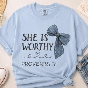 May include: A light blue short-sleeve t-shirt featuring the black text "SHE IS WORTHY" above a small heart graphic and the text "PROVERBS 31". A large, dark blue denim-textured bow graphic is positioned to the right of the text on the shirt.