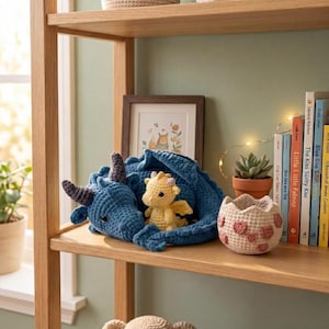 May include: A shelf displays a blue crocheted dragon with gray horns, cradling a yellow dragon. A crocheted egg-shaped basket with pink hearts sits nearby. A framed picture and books are also on the shelf.