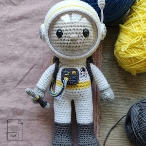 May include: A crocheted astronaut doll featuring a white suit with yellow accents, dark gray boots, and a clear plastic helmet visor. The figure includes a dark gray chest pack with a blue hose, a small embroidered astronaut patch, and a handheld tool.