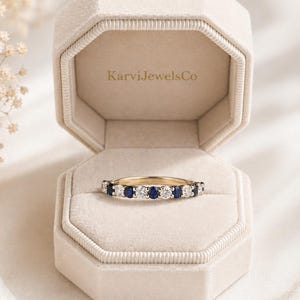 May include: A gold band ring with alternating round white and blue gemstones, displayed in an open, octagonal, beige velvet jewelry box. The box has the text "KarviJewelsCo" on the inside. The ring is set against a soft, neutral background.