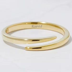 Peut inclure: Une bague dorée avec un design ouvert et enveloppant. La bague a une surface lisse et polie et porte le texte "Karvij". Le design de la bague est moderne et minimaliste, avec une courbe subtile.