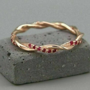 Könnte beinhalten: Ein roségoldener Ring mit einem gedrehten Design, verziert mit kleinen roten Edelsteinen. Der Ring liegt auf einer grauen, strukturierten Oberfläche und zeigt seine komplizierten Details und seinen eleganten Stil. Das Design des Rings ist eine durchgehende Schleife.