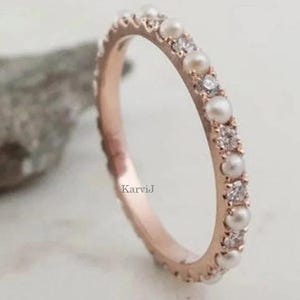 Peut inclure: Une bague en or rose ornée d'un motif alterné de petites perles blanches et de pierres rondes transparentes serties sur la circonférence extérieure. Le mot KarviJ est imprimé en petites lettres noires sur la surface intérieure de l'anneau.