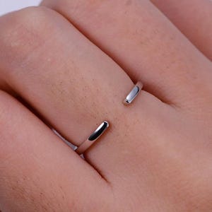 Puede incluir: Un anillo abierto de color plateado. El anillo es una banda simple con un espacio, creando un diseño ajustable. El anillo se lleva en un dedo, mostrando su estilo minimalista y su estética moderna. La superficie pulida del anillo refleja la luz.