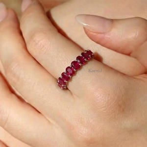 Op de afbeelding: Een zilveren ring met een reeks ovaal geslepen, dieprode edelstenen. De edelstenen zijn dicht op elkaar geplaatst, waardoor een doorlopende band van kleur ontstaat. De ring wordt getoond op een vinger, waarbij het ontwerp en de levendige rode tint van de stenen worden benadrukt.