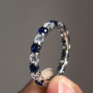 Può includere: Una fede a giro completo color argento con un motivo alternato di zaffiri blu scuro e diamanti bianchi trasparenti a taglio rotondo, fissati con una montatura a griffe condivisa che circonda l'intera circonferenza dell'anello.
