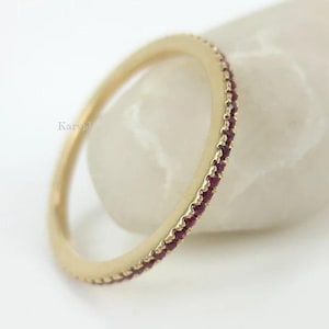 Op de afbeelding: Een dunne gouden ring met een doorlopende rij kleine, ronde, donkerrode edelstenen langs de buitenrand. Het metaal heeft een gladde, gepolijste afwerking en de ring is in een hoek geplaatst om de cirkelvorm en de stenen goed te tonen.