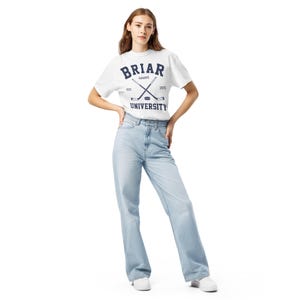 May include: A white short-sleeve t-shirt featuring the dark blue text BRIAR UNIVERSITY, HAWKS, EST. 2015, and a graphic of two crossed hockey sticks with a puck, paired with light blue denim jeans and white sneakers.