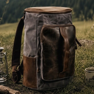 May include: A cylindrical backpack constructed from grey canvas and brown leather. It features a large rectangular front pocket with a vertical leather handle, a top lid, and adjustable shoulder straps with metal hardware accents.