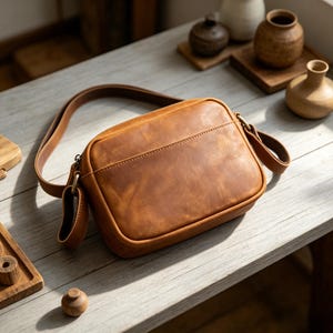 Puede incluir: Bolso bandolera de cuero marr&oacute;n con una correa larga. El bolso tiene forma rectangular con esquinas redondeadas y un bolsillo frontal. El cuero tiene un aspecto envejecido. El bolso est&aacute; sobre una superficie de madera clara.