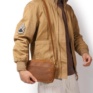 Puede incluir: Un bolso bandolera de cuero marr&oacute;n con forma rectangular y una correa larga y ajustable. La persona lleva una chaqueta color beige con un parche de estilo militar en la manga. El bolso tiene un dise&ntilde;o sencillo y cl&aacute;sico.